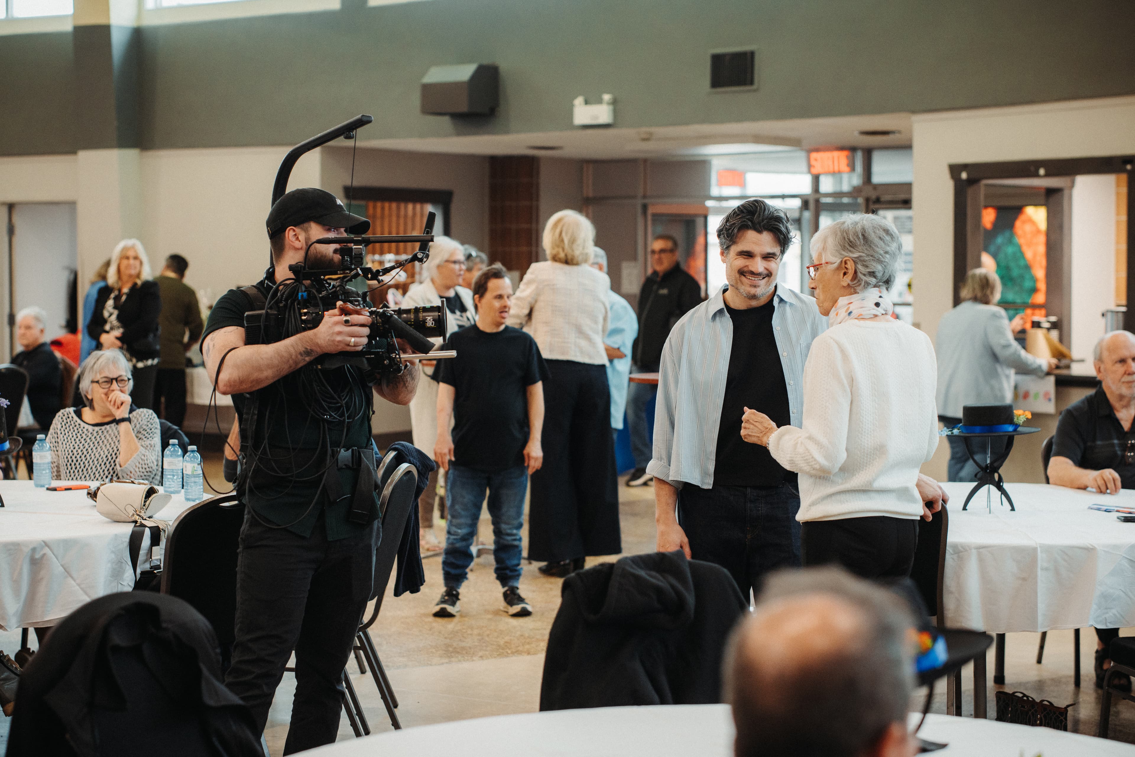 Tournage dans une salle communautaire : une intervenante discute avec une participante pendant qu'une caméra capte la scène, entourées de membres du groupe